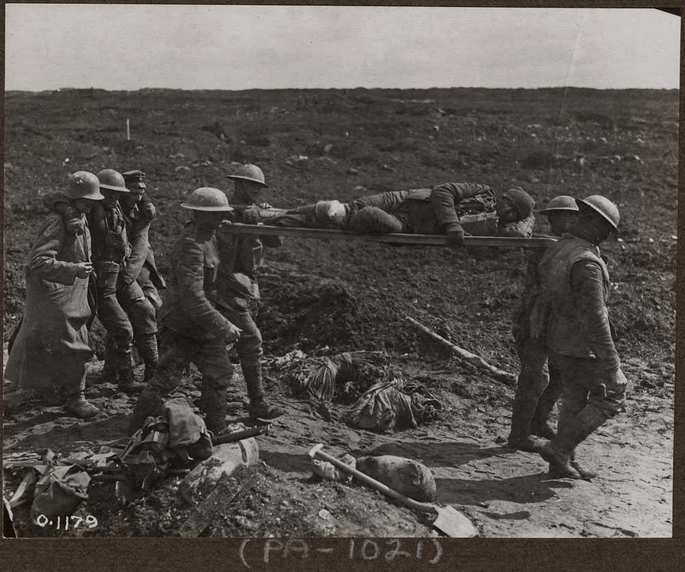 Photographie en noir et blanc – Un champ de bataille boueux. Quatre hommes en uniforme militaire et portant des casques transportent sur leurs épaules un homme allongé sur une civière. Trois hommes les suivent, celui au centre soutenu par les deux autres.